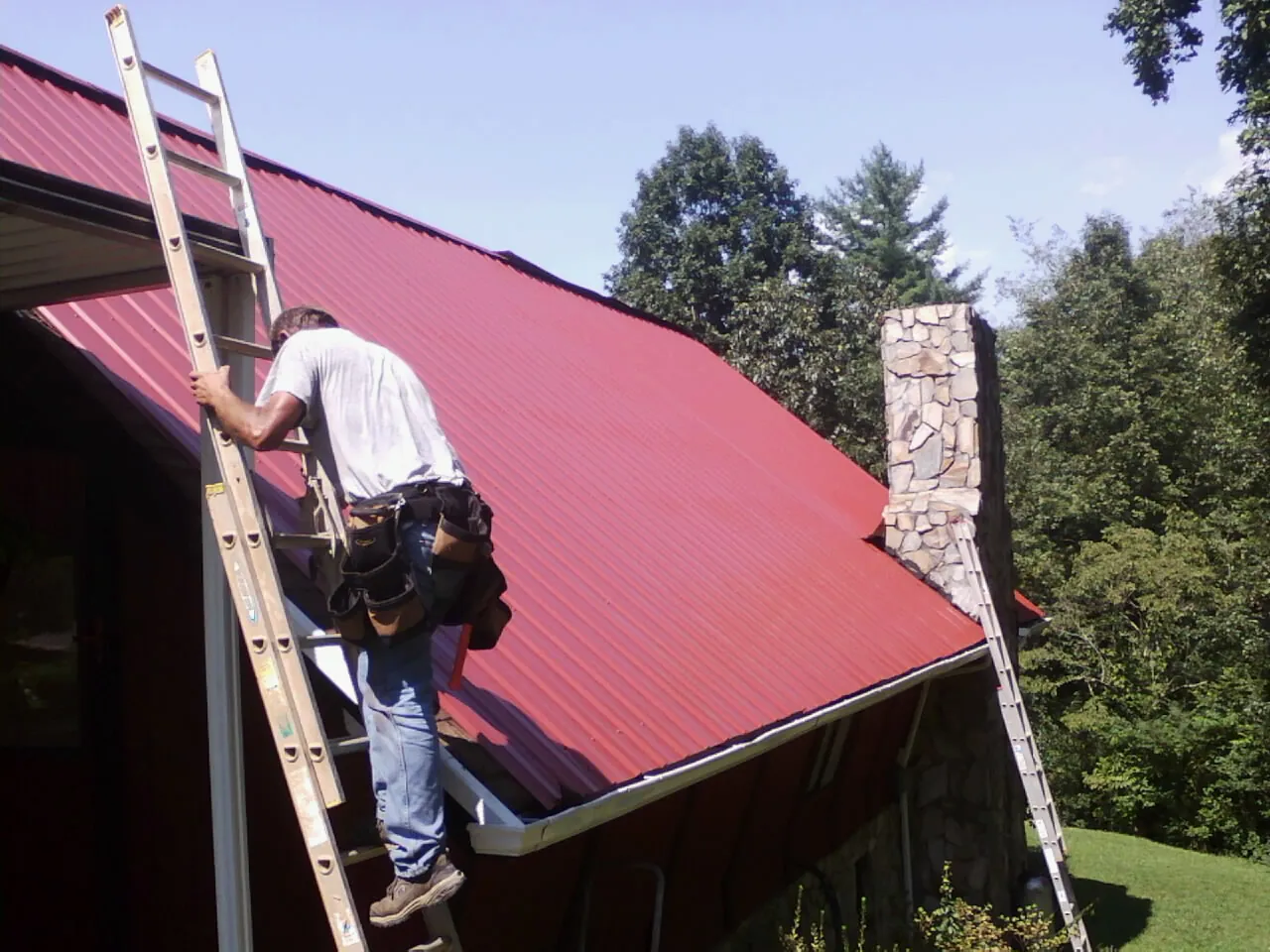 Roofer on a ladder beside a red metal roof and stone chimney — Soffit & Fascia Repair by Roofing Brothers in Skaneateles